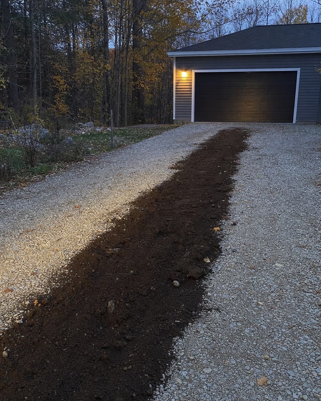 Une tranchée rebouchée traversant une allée de gravier vers un garage détaché, borne allumée au mur